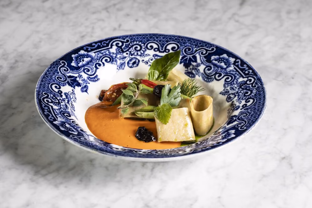 Blue and white decorative bowl on a marble surface, containing a gourmet dish with orange sauce, assorted vegetables, herbs, and small pasta pieces.