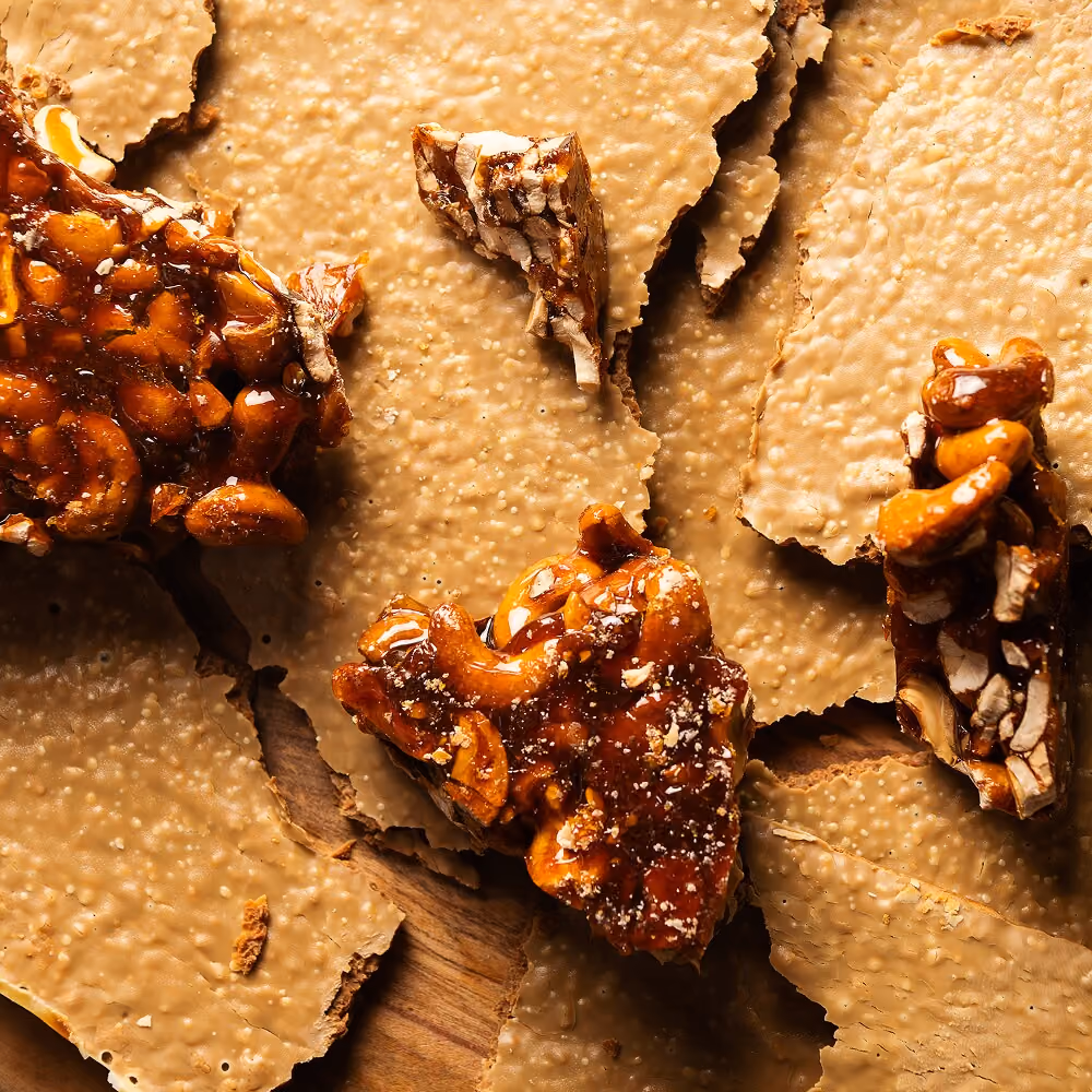 Close-up of broken pieces of peanut brittle with glossy caramel-coated nuts on top of lighter-colored brittle pieces on a wooden surface.